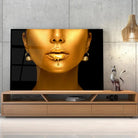 Tableau Femme maquillage en or en verre acrylique, posé sur un meuble TV en bois devant un mur en béton