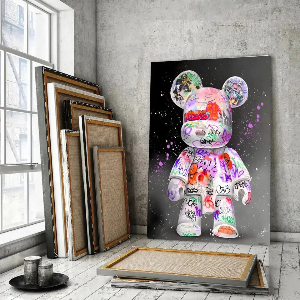 Tableau Street Art Kaws Bearbrick sur toile, posé devant des cadres empilés près d'une fenêtre
